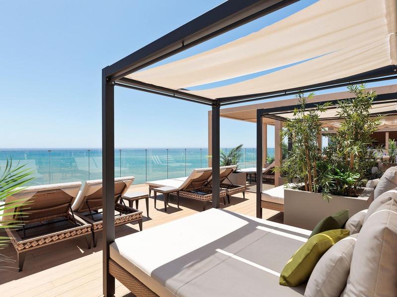 Moderner Balkon mit Holzboden, weißen Liegen, Pflanzen und Blick aufs blaue Meer und Himmel.