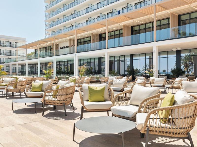 Moderne Hotelterrasse mit geflochtenen Stühlen, kleinen Tischen und Glasfassade im Hintergrund.