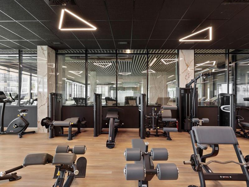 Moderne Fitnessstudio mit Geräten, Holzfußboden und Glaswänden, dreieckige Deckenlichter.