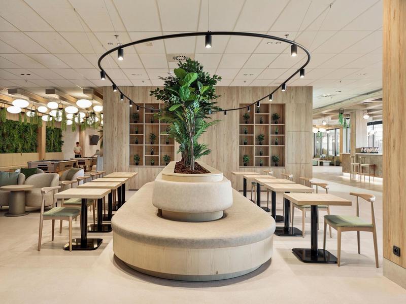 Moderner Gastraum mit runden Sitzbänken, zentralem Baum und hellen Holztischen.