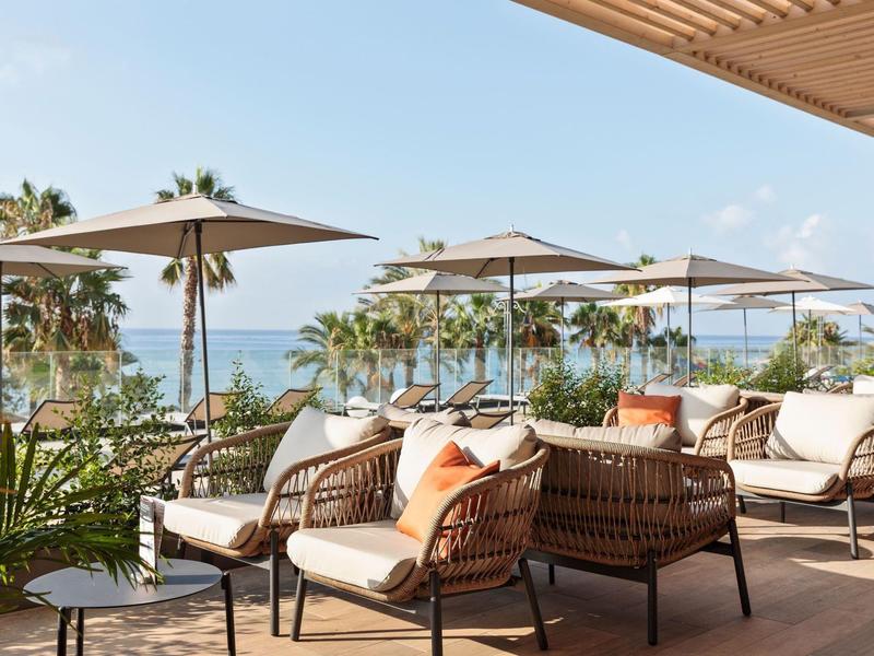 Outdoor-Lounge mit Korbstühlen und Sonnenschirmen, Blick auf Meer und Palmen bei strahlendem Himmel.