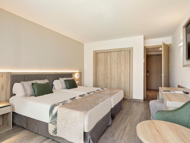 Helles Hotelzimmer mit zwei Betten, Holzschrank, grünem Sessel und Flachbildfernseher an der Wand.