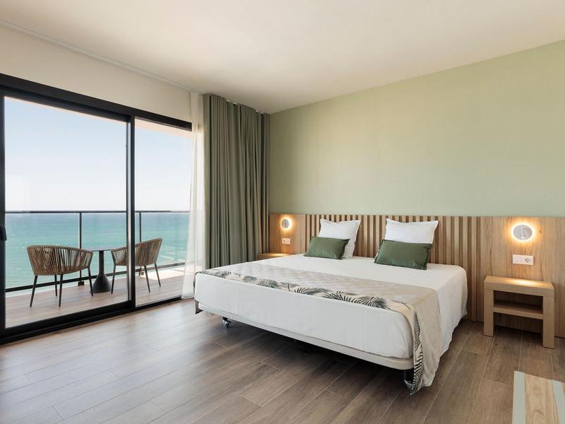 Helles Schlafzimmer mit Doppelbett, Holzlamellenwand, grüner Wandfarbe und Balkon mit Meerblick.