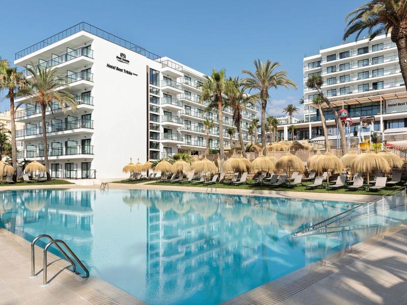 Großer blauer Pool vor weißen Hotelgebäuden mit Palmen und Liegestühlen im Sonnenlicht.
