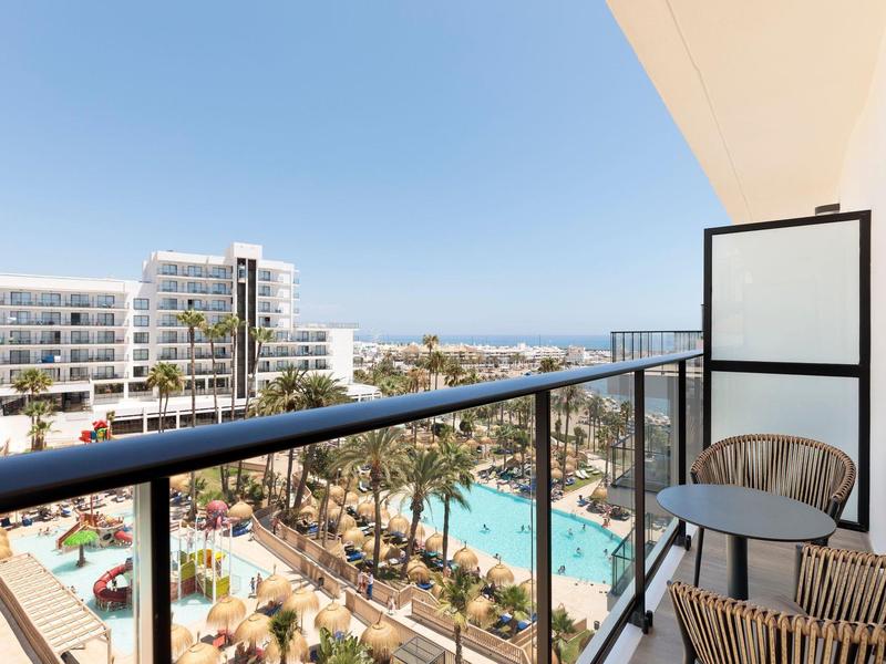 Balkon mit Stuhl und Tisch, Blick auf Pool, Palmen und moderne weiße Gebäude bei blauem Himmel.
