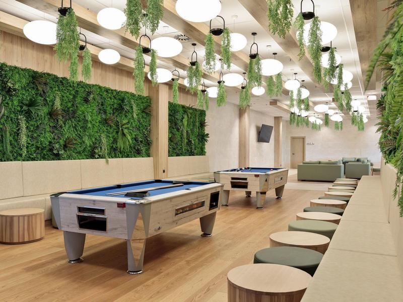 Moderner Spielraum mit Billardtischen, hängenden Pflanzen und beige Sitzbänken mit Holzhockern.