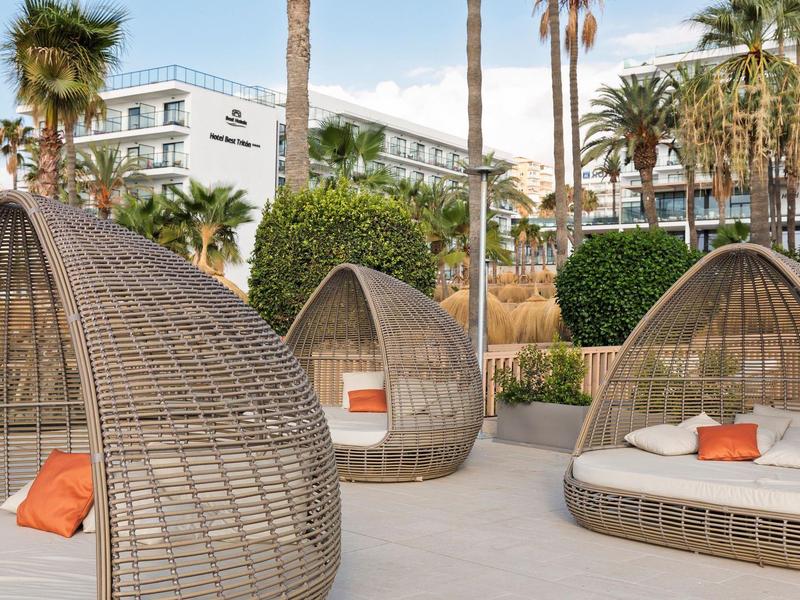 Outdoor-Loungemöbel aus geflochtenem Material mit weißen Polstern und orangen Kissen vor Hotel.