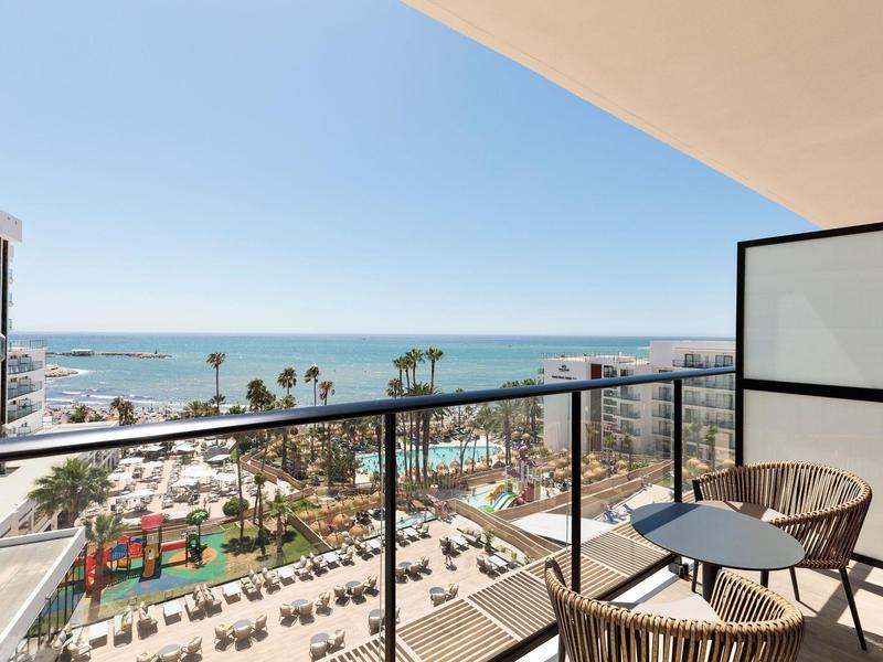 Balkon mit zwei Stühlen und Tisch, Blick auf Pool, Palmen und Meer unter klarem Himmel.