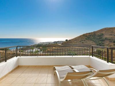 Balkon mit zwei Liegestühlen, Geländer und Aussicht auf Meer und hügelige Landschaft.