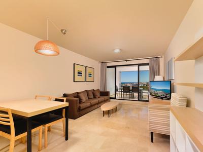 Helles Wohnzimmer mit Sofa, Esstisch, Fernseher und Balkon mit Meerblick bei Sonnenuntergang.