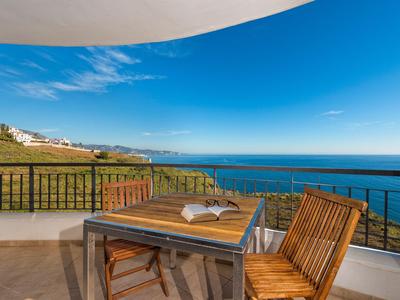 Balkon mit Holztisch und Stühlen, offenes Buch, Meerblick unter blauem Himmel.