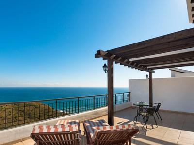 Terrassenbereich mit Holzgitter, Tisch, Stühlen und Meerblick unter klarem blauem Himmel.