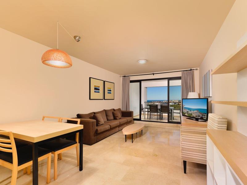 Helles Wohnzimmer mit Sofa, Esstisch, Fernseher und Balkon mit Meerblick bei Sonnenuntergang.