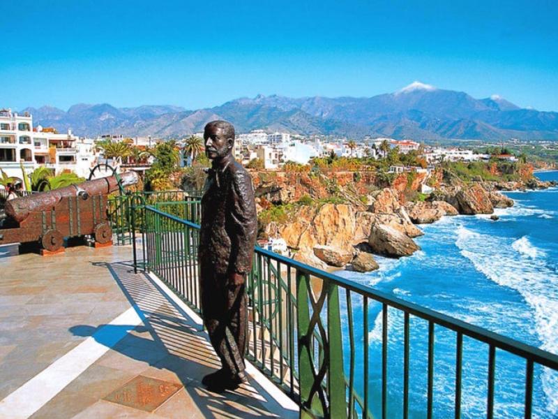 Statue eines Mannes an Geländer mit Blick auf felsige Küste und blaues Meer bei klarem Himmel.