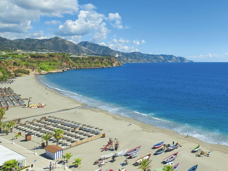 Breiter Sandstrand mit Liegestühlen, bunten Kajaks und bewaldeten Bergen am Horizont.