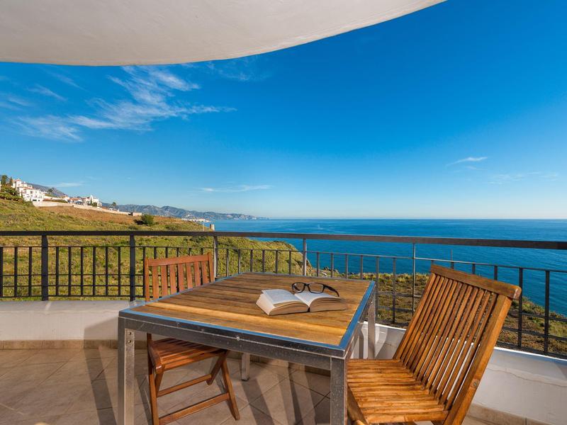 Balkon mit Holztisch und Stühlen, offenes Buch, Meerblick unter blauem Himmel.