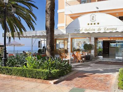 Eingangsbereich eines Hotels mit Terrasse, Palmen, Pflanzen und Blick auf das Meer im Hintergrund.
