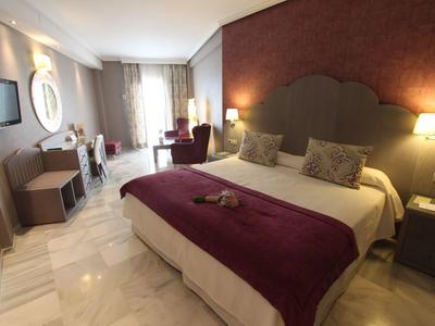 Modernes Hotelzimmer mit großem Doppelbett, violettem Überwurf, Schreibtisch, Fernseher und bequemen Sesseln.