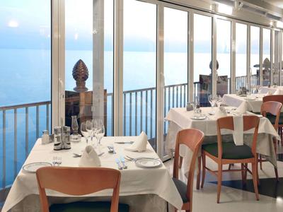 Hell eingerichtetes Restaurant mit weißen Tischen und Holzstühlen, großer Fensterfront mit Meerblick.