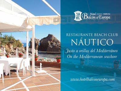 Terrasse mit weißen Tischen und Stühlen, Meerblick, Felsen, Sonnensegel, blau-weiße Farbgebung.