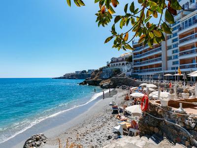 Strand mit Sand und Kies, blaues Meer, klare Himmel, Sonnenschirme und Gebäude am Ufer.