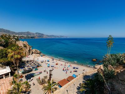 Sandstrand mit bunten Sonnenschirmen, klarem blauem Meer und Bergen im Hintergrund bei sonnigem Himmel.