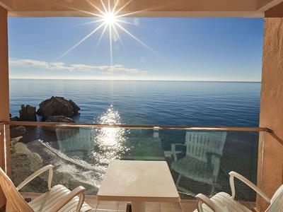 Balcón con mesa y sillas con vista al mar tranquilo y el sol en un cielo despejado.