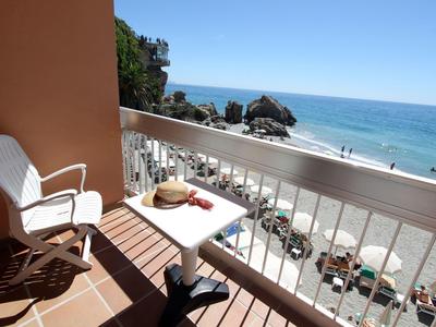 Balkon mit weißem Stuhl, Tisch, Sonnenhut und Blick auf Sandstrand, Felsen und blaues Meer.
