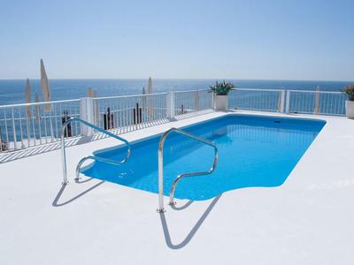 Blauer Pool auf weißer Terrasse mit Meerblick und bewölktem Himmel im Hintergrund.