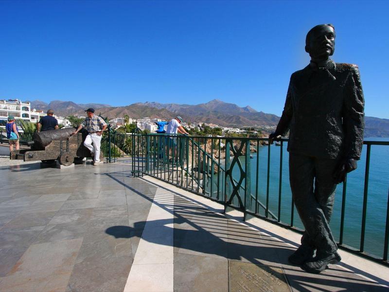 Promenade mit dunkler Bronzestatue eines Mannes, blauem Himmel und Bergen im Hintergrund.