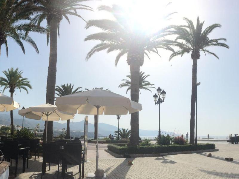 Strandpromenade mit Palmen, Sonnenschirmen und Tischen bei klarem Himmel und Sonnenlicht.