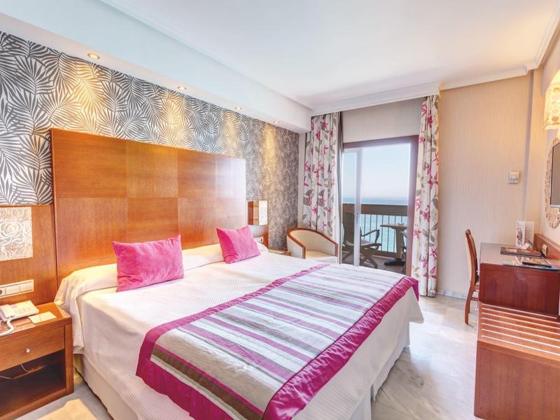 Helles, modernes Schlafzimmer mit großem Doppelbett, bunten Kissen und Zugang zu Balkon mit Meerblick.