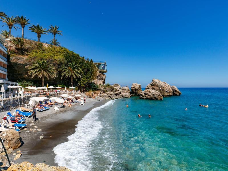 Strand mit dunklem Sand, türkisfarbenem Wasser, Palmen und Liegestühlen unter blauem Himmel.
