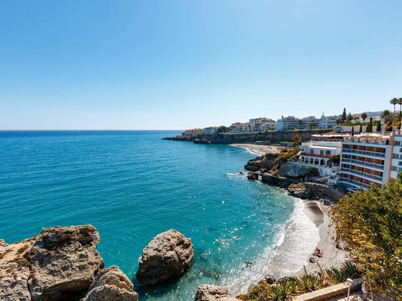 Kleine Sandbucht mit Felsen, blauem Meer und Gebäuden am Küstenhang unter klarem Himmel.