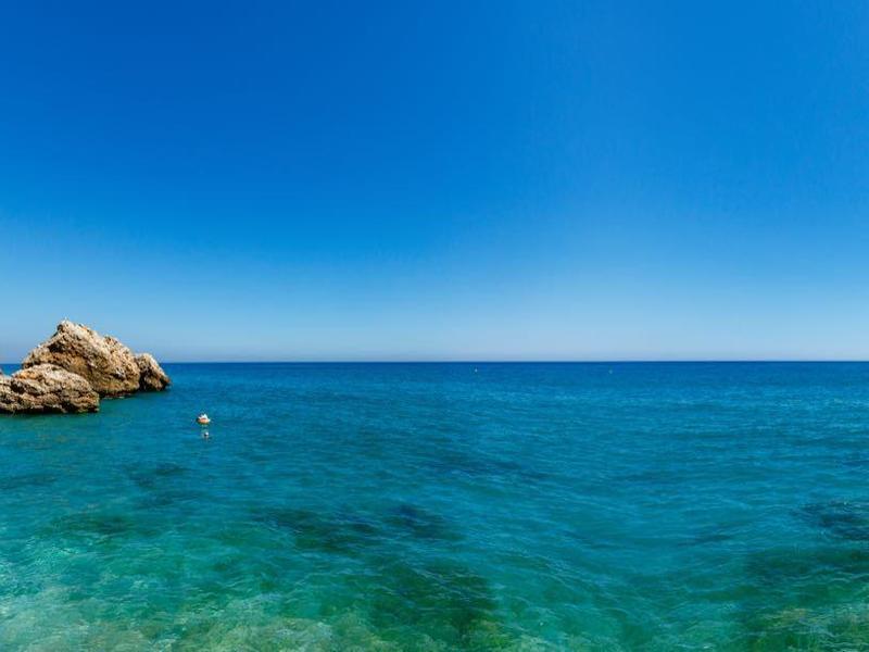 Klarer blauer Himmel über ruhigem türkisfarbenem Meer mit Felsen und wenigen Menschen am Strand.