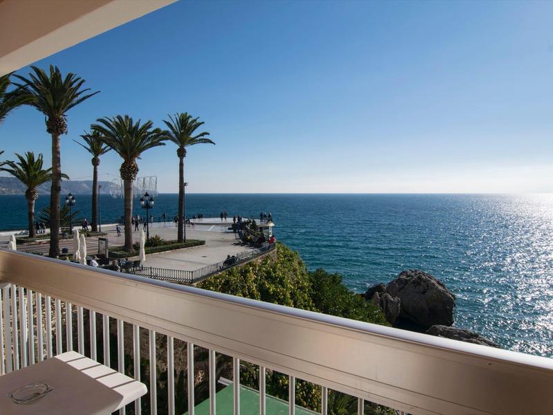 Balkon mit weißem Geländer, Palmen, blauem Meer und klarem Himmel im Hintergrund am sonnigen Tag.