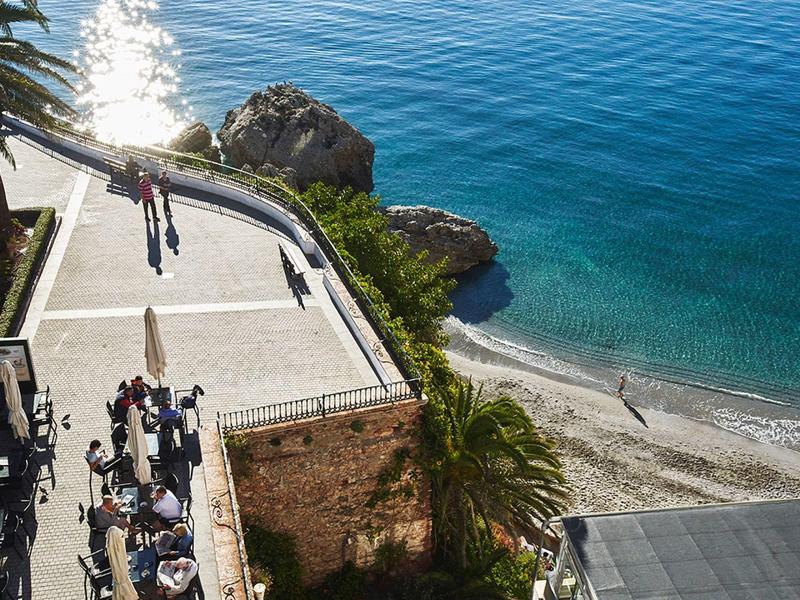 Menschen am sonnigen Strand mit klarem blauem Wasser und felsigem Uferabschnitt.