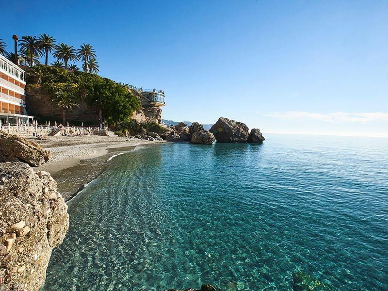 Klares, ruhiges Meer vor felsiger Küste mit Palmen, Strand und Gebäuden unter blauem Himmel.