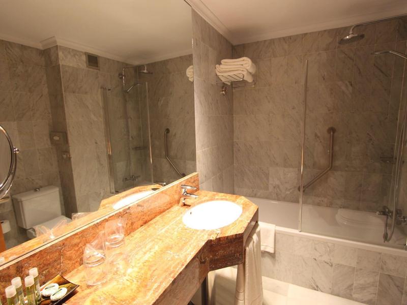 Hell beleuchtetes Badezimmer mit Marmorwaschtisch, rundem Spiegel und Badewanne mit Glaswand.