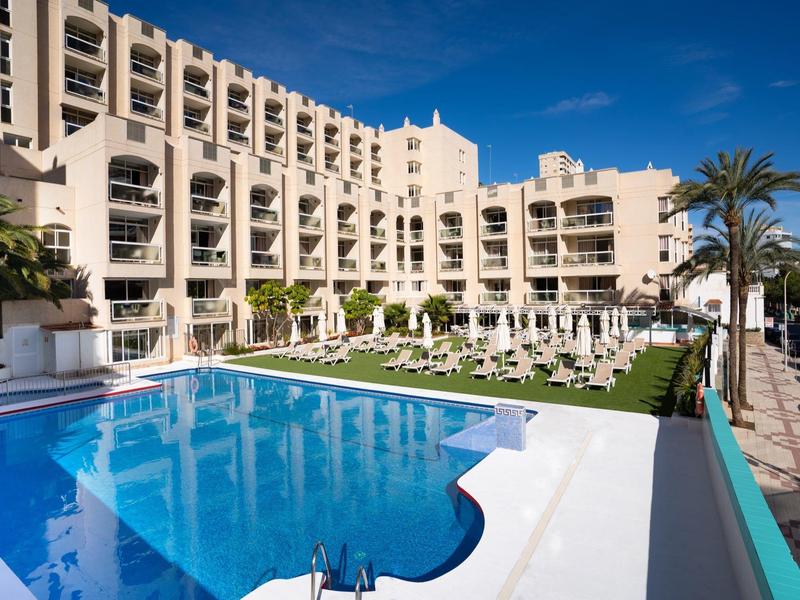 Modernes Hotel mit großem Pool, Liegestühlen und Palmen bei blauem Himmel.