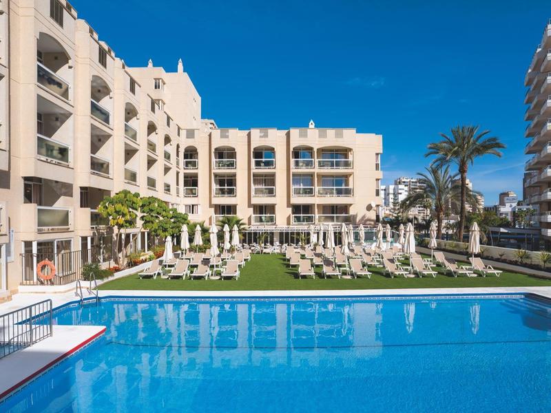 Moderner Hotelpool mit Liegestühlen und Palmen vor einem mehrstöckigen Gebäude bei klarem Himmel.