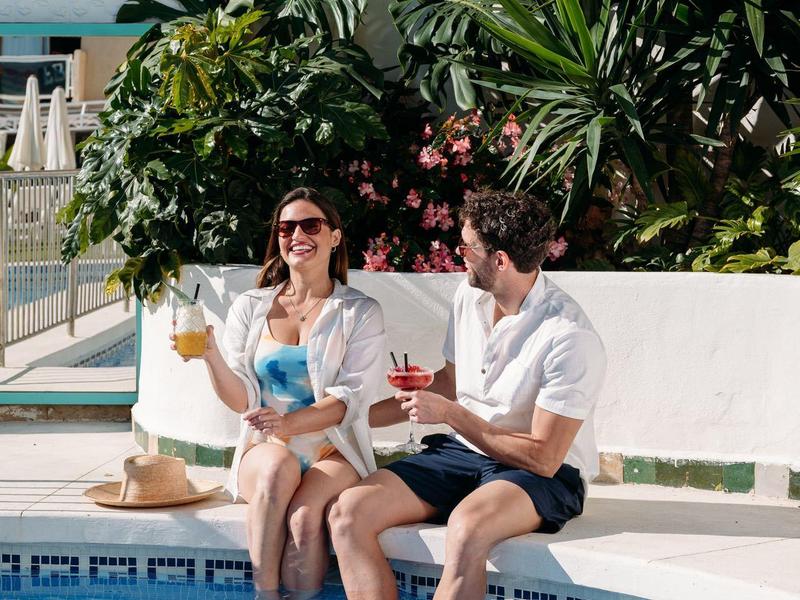 Paar entspannt am Poolrand mit Getränken, umgeben von tropischer Vegetation.