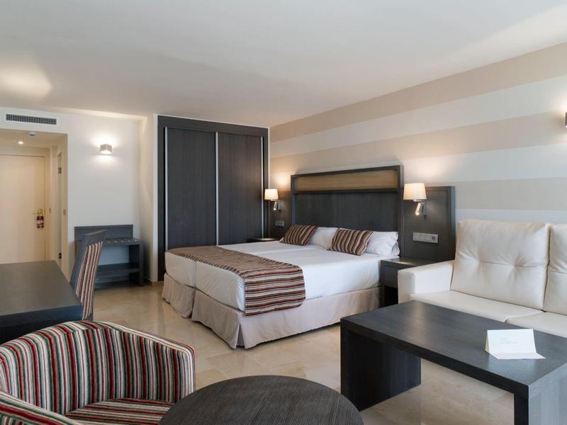 Helles Hotelzimmer mit Doppelbett, Sessel, Schreibtisch und Flachbildfernseher an der Wand.