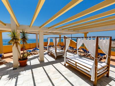 Terrasse mit Holzpergola, Liegen mit weißen Vorhängen, blauer Himmel und Meer im Hintergrund.