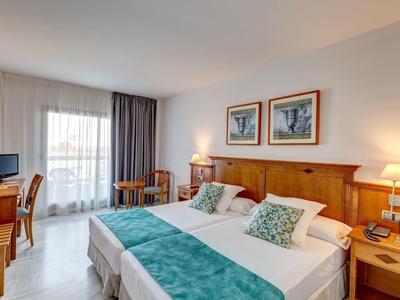 Helles Hotelzimmer mit zwei Betten, Holzmöbeln, grünen Akzenten und Balkonfenster mit Vorhängen.