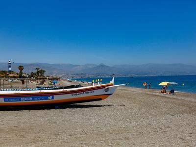 Ein bunter Fischerboot liegt am Sandstrand, blauer Himmel und ruhiges Meer mit Menschen im Hintergrund.