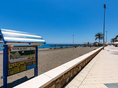 Strandpromenade mit Wegweiser, Sandstrand, blauer Himmel und Palmen am Meer.