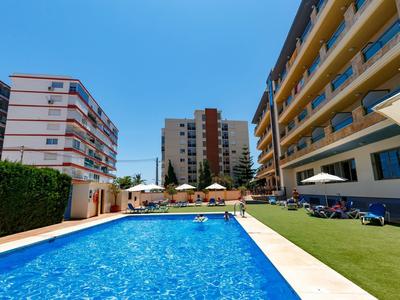 Schwimmbad mit blauem Wasser, umgeben von Liegestühlen und mehrstöckigen Wohngebäuden bei Sonnenschein.