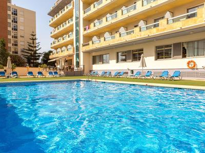 Schwimmbad mit klarem Wasser vor gelbem Gebäudekomplex und blauen Liegestühlen.