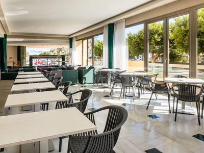 Helles Café mit weißen Tischen, schwarzen Stühlen und großen Fenstern zum Außenbereich mit Bäumen.